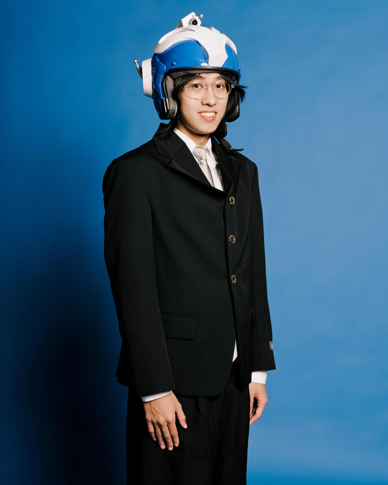 Allex Chan, standing against a blue backdrop in a black suit and white shirt, wearing a white and blue flight helmet and round glasses.